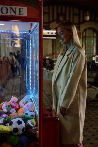 Smiling woman in a beige coat playing a claw machine game in an amusement arcade.
